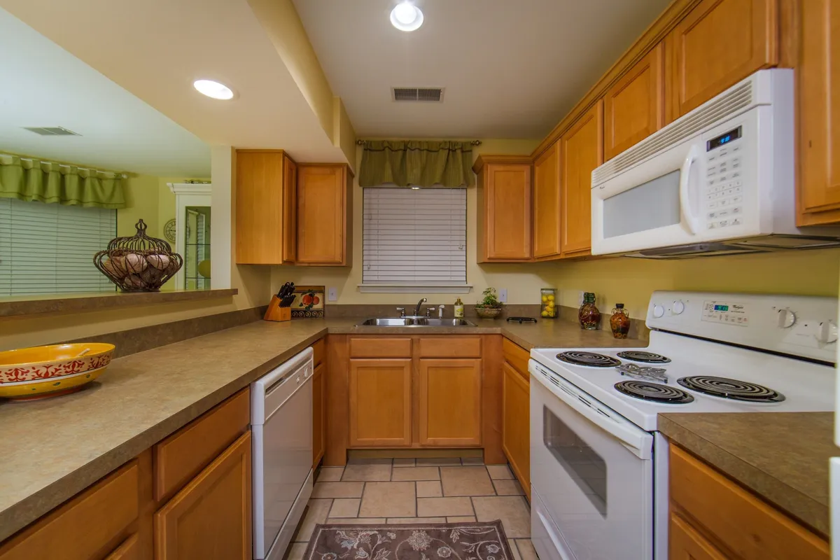 This image displays a welcoming kitchen with wooden cabinets, modern appliances—including a full-sized refrigerator and microwave—and a decorative bowl on the counter. Sunlight filters in through a curtained window, enhancing the cozy, usable feel of the space. The layout invites group cooking sessions and casual conversations, perfect for families or friends gathering for meals.