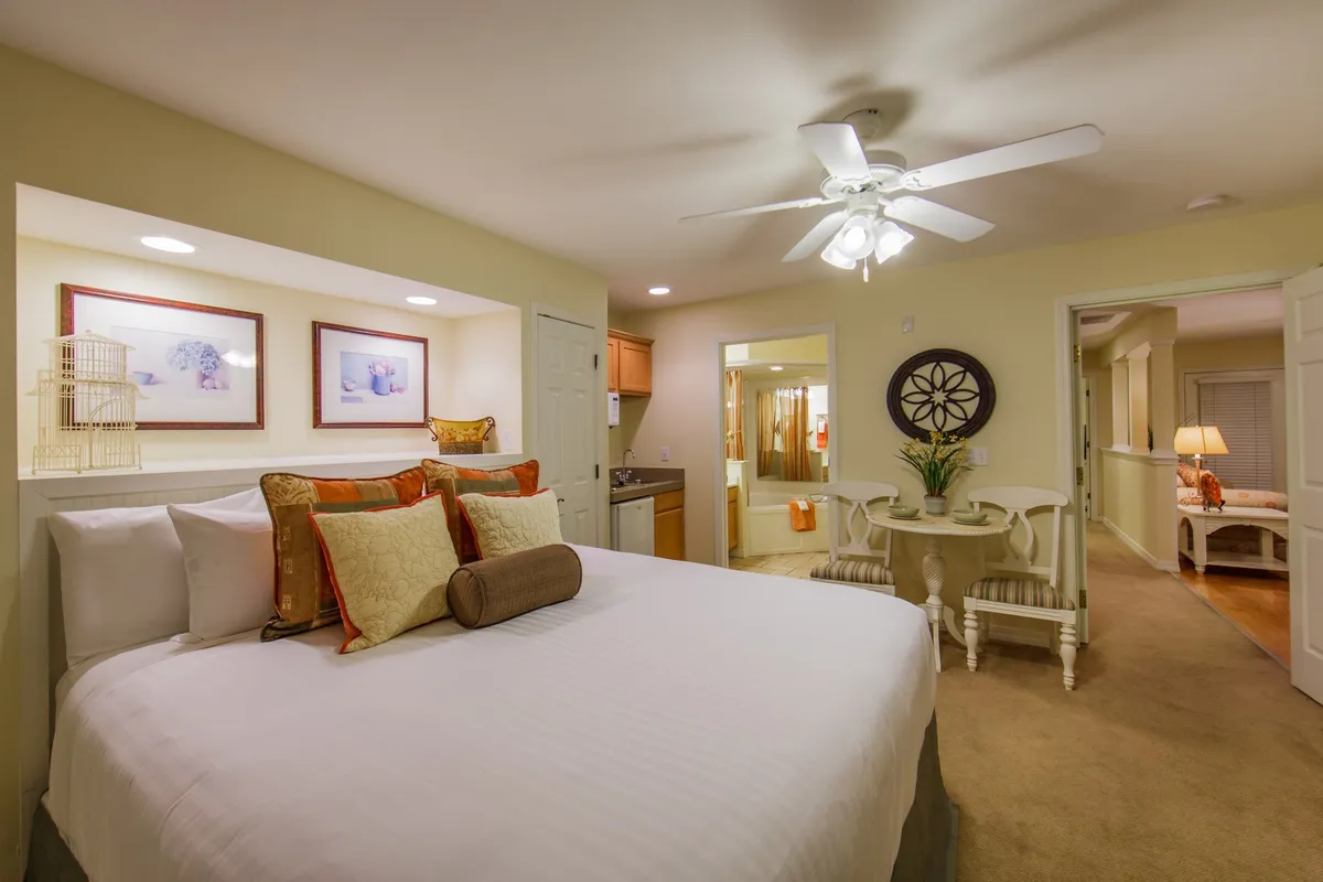 A warmly lit bedroom featuring a plush bed adorned with decorative pillows, a dining table set for two, and tasteful wall art. The scene conveys a sense of welcoming comfort and intimate relaxation, ideal for unwinding after a day at the resort.