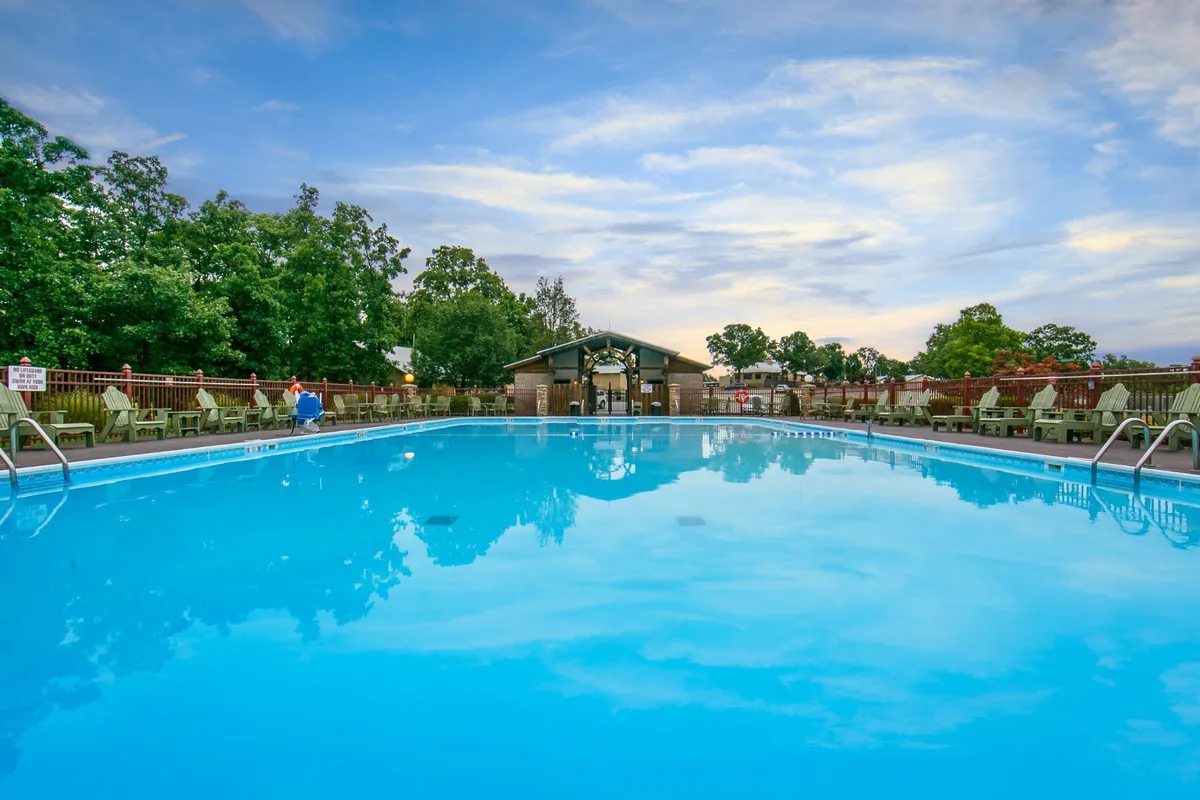 An inviting outdoor swimming pool is surrounded by lush trees, ample poolside seating, and a pavilion for shade. The scene suggests both daytime relaxation and lively social gatherings, with sunset reflections enhancing the vibrant, communal energy.