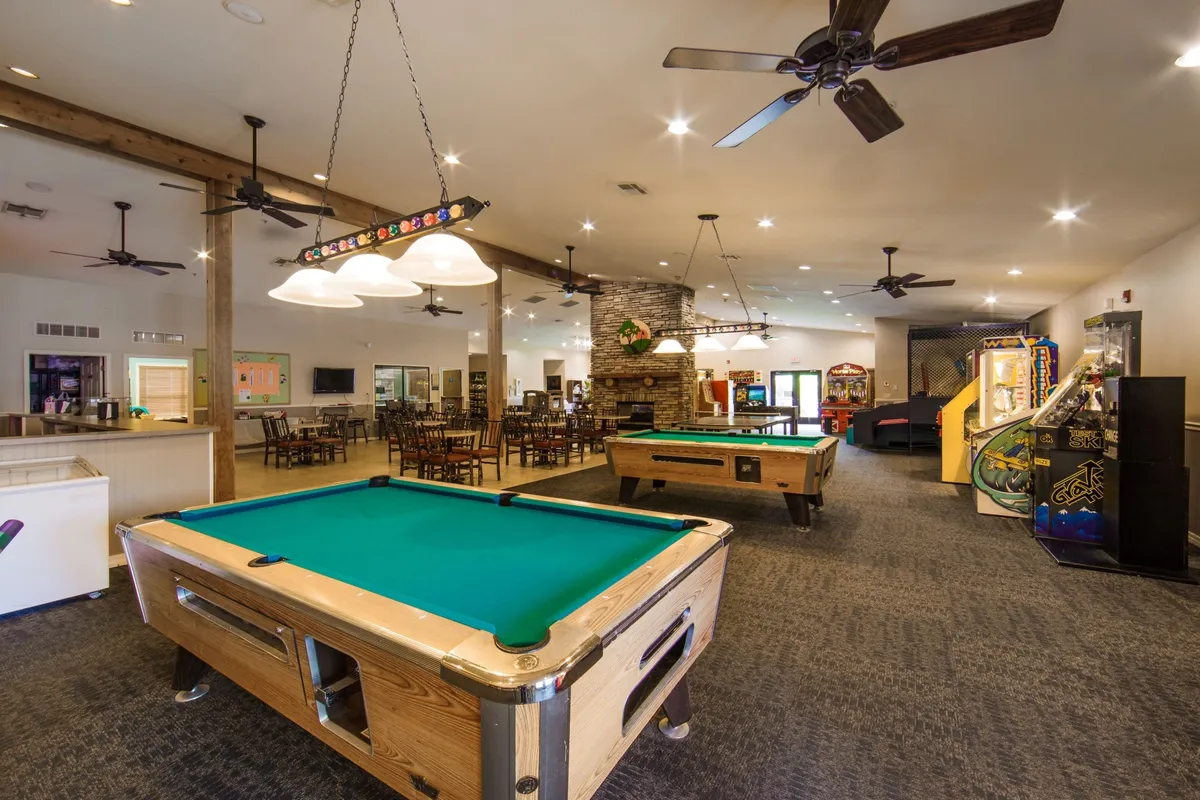 A spacious entertainment area that includes a dining space adjacent to pool tables and a stone fireplace. The dining area is set for socializing and celebratory meals, framed by elements of fun—like arcade games—suggesting a lively, multi-purpose venue for group gatherings and shared enjoyment.