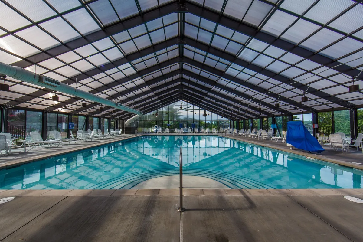 This image features a spacious indoor swimming pool with a striking glass roof, allowing natural light to pour in while offering year-round enjoyment. Lounge chairs are arranged along the poolside, with gentle reflections shimmering on the water's surface, evoking a tranquil and inviting atmosphere.
